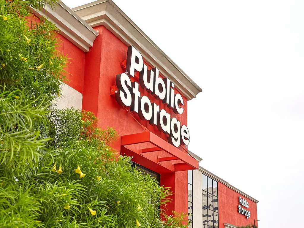 Exterior view of a Public Storage facility with a bright red facade and the company's logo prominently displayed. Green foliage partially obscures the lower left corner of the building.