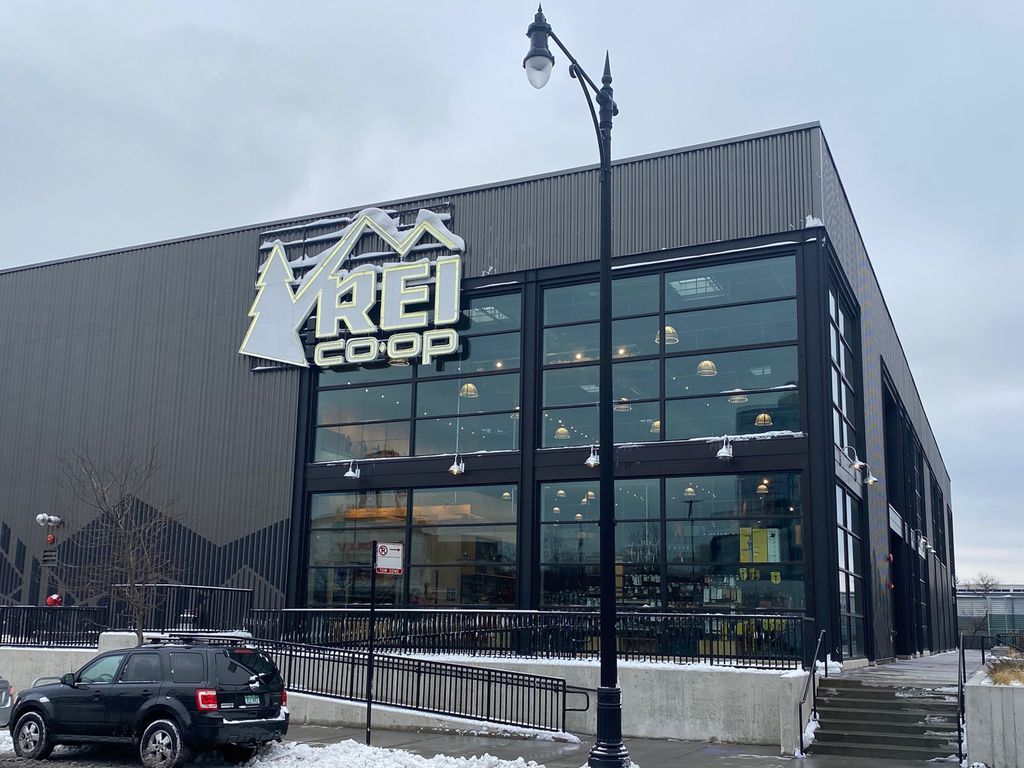 Exterior view of an REI Co-op store, featuring the company logo on a gray building with large windows. A black SUV is parked in front, and a street lamp stands nearby.