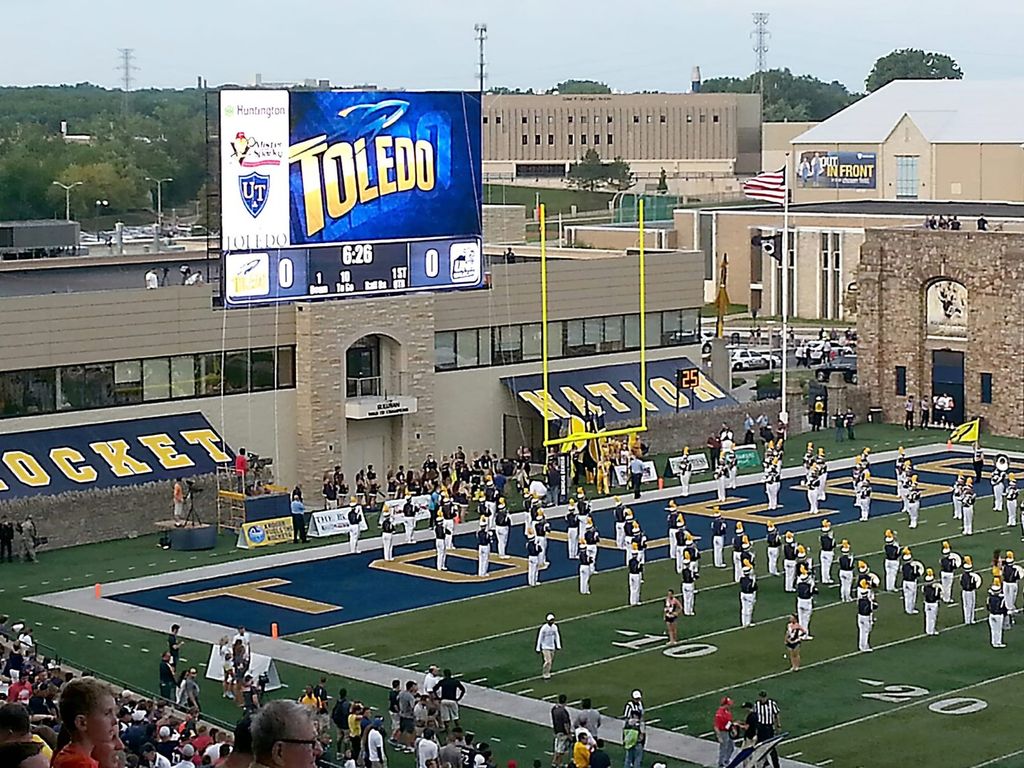 A view of a Toledo Rockets football game with the marching band performing on the field. The scoreboard shows the Toledo logo and game information.